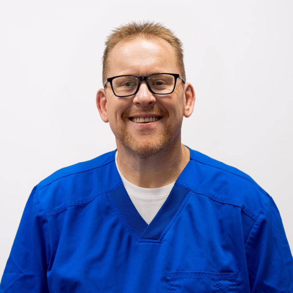 Smiling man in blue scrubs and glasses, looking at the camera.