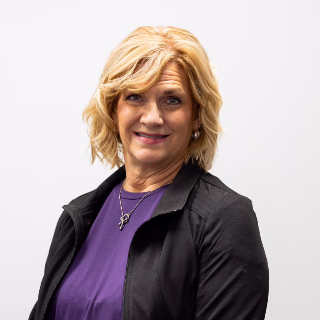 Portrait of a smiling woman with short blonde hair, wearing a purple blouse and black jacket against a light background.